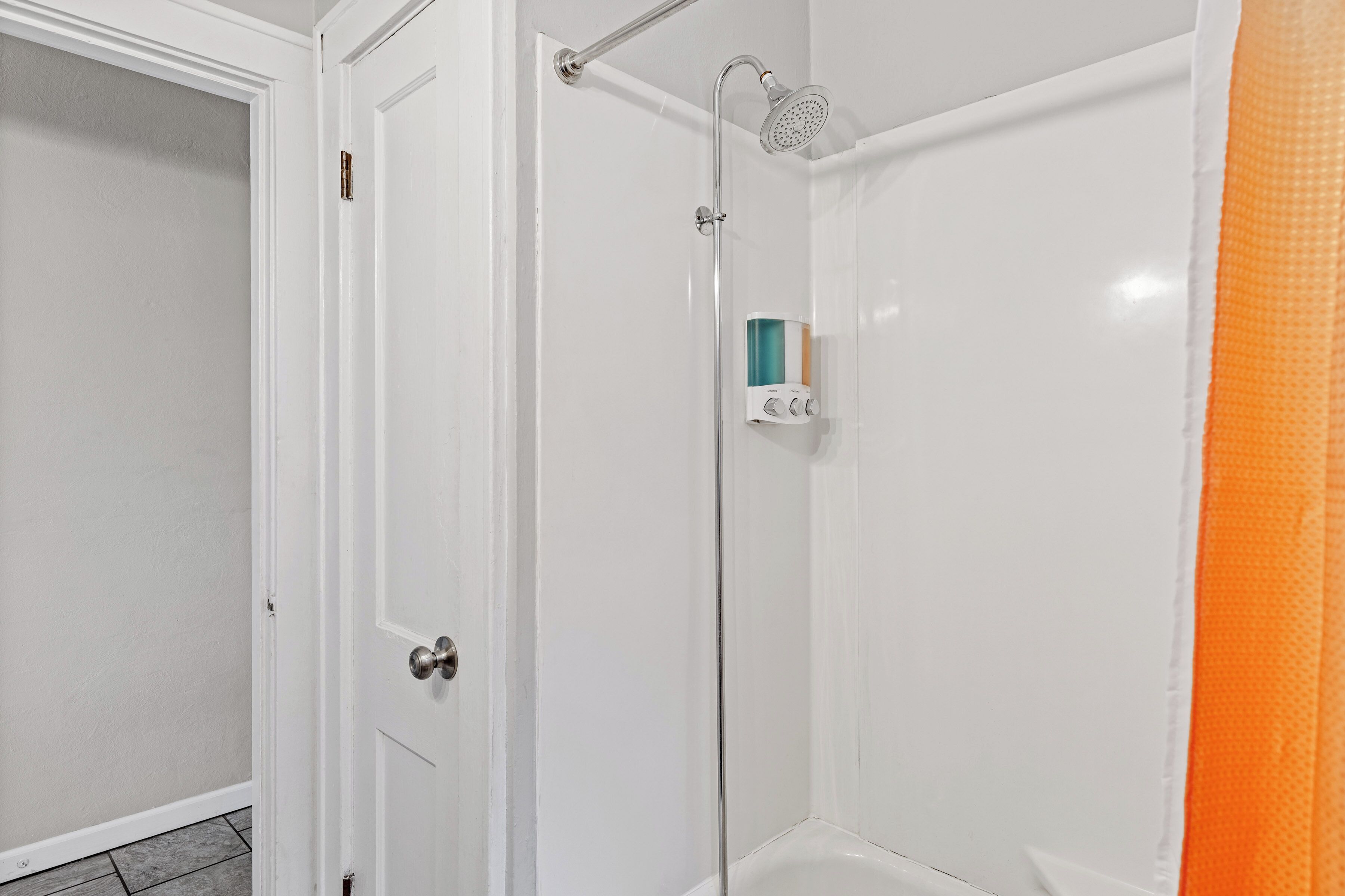 Ein Badezimmer mit einer weißen Tür, einer Dusche inklusive Handbrause und einem Seifenspender an der Wand.