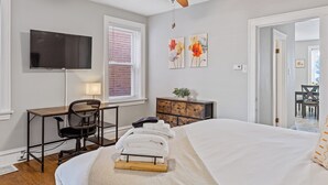 A bedroom with a bed, a desk, a chair, a TV, and artwork on the wall.