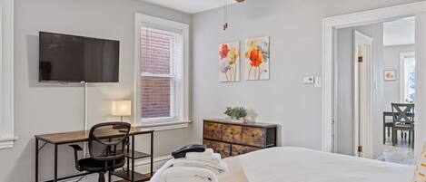 A bedroom with a bed, a desk, a chair, a TV, and artwork on the wall.