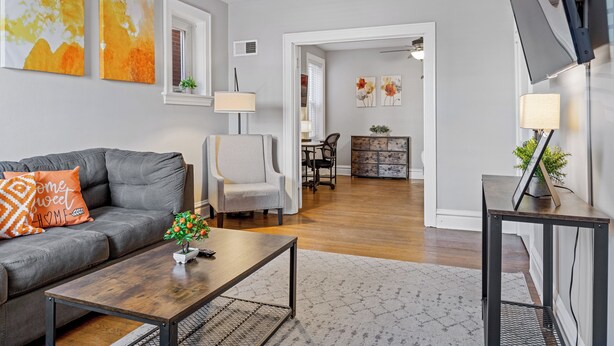 A modern living room with a grey sofa, a coffee table, and a TV on a stand. There is a dining area with a table and chairs in the background.