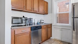 A kitchen with wooden cabinets, a stainless steel refrigerator, a dishwasher, a microwave, and a window with blinds.