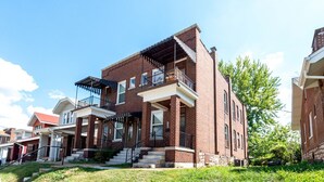 A two-story brick building with a balcony on the second floor, a front porch, and a small front yard.