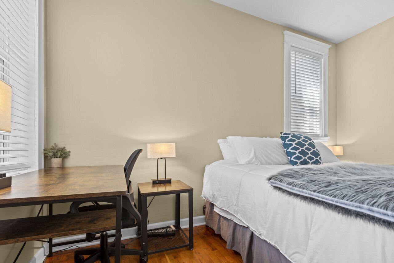A bedroom with a bed, a desk, a chair, and a small table with a lamp.
