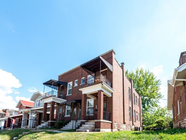 A two-story brick building with a balcony on the second floor, a front porch, and a small front yard.