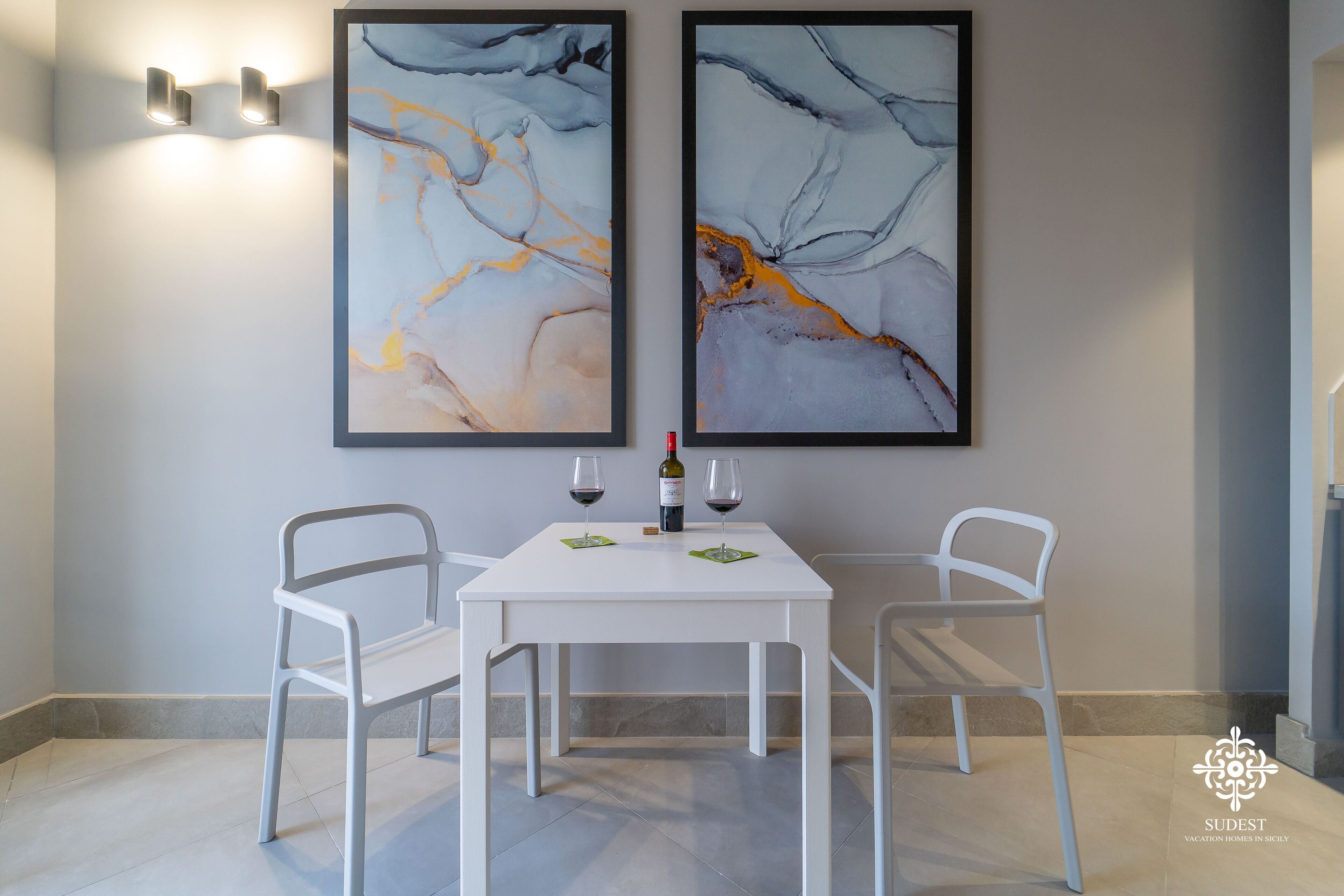 Un coin repas avec une table et des chaises blanches, deux tableaux abstraits accrochés au mur et une bouteille de vin.