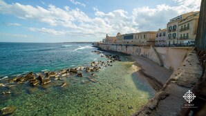 A coastal view with clear waters, a rocky shoreline, and a row of buildings along the sea. - Matteotti Luxury Residence (Syracuse)
