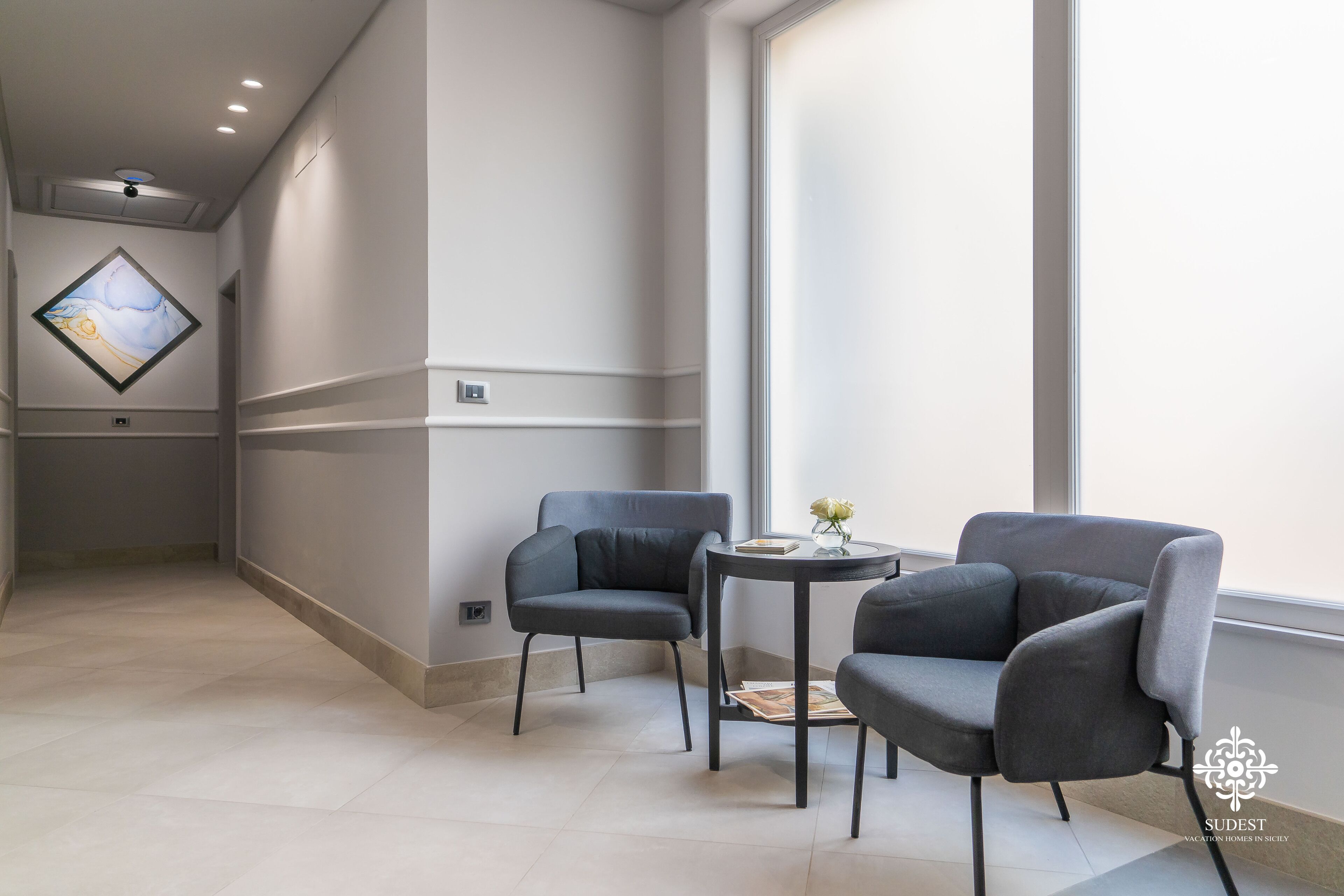 A modern hotel room with two grey armchairs, a small round table, and a large window providing natural light.