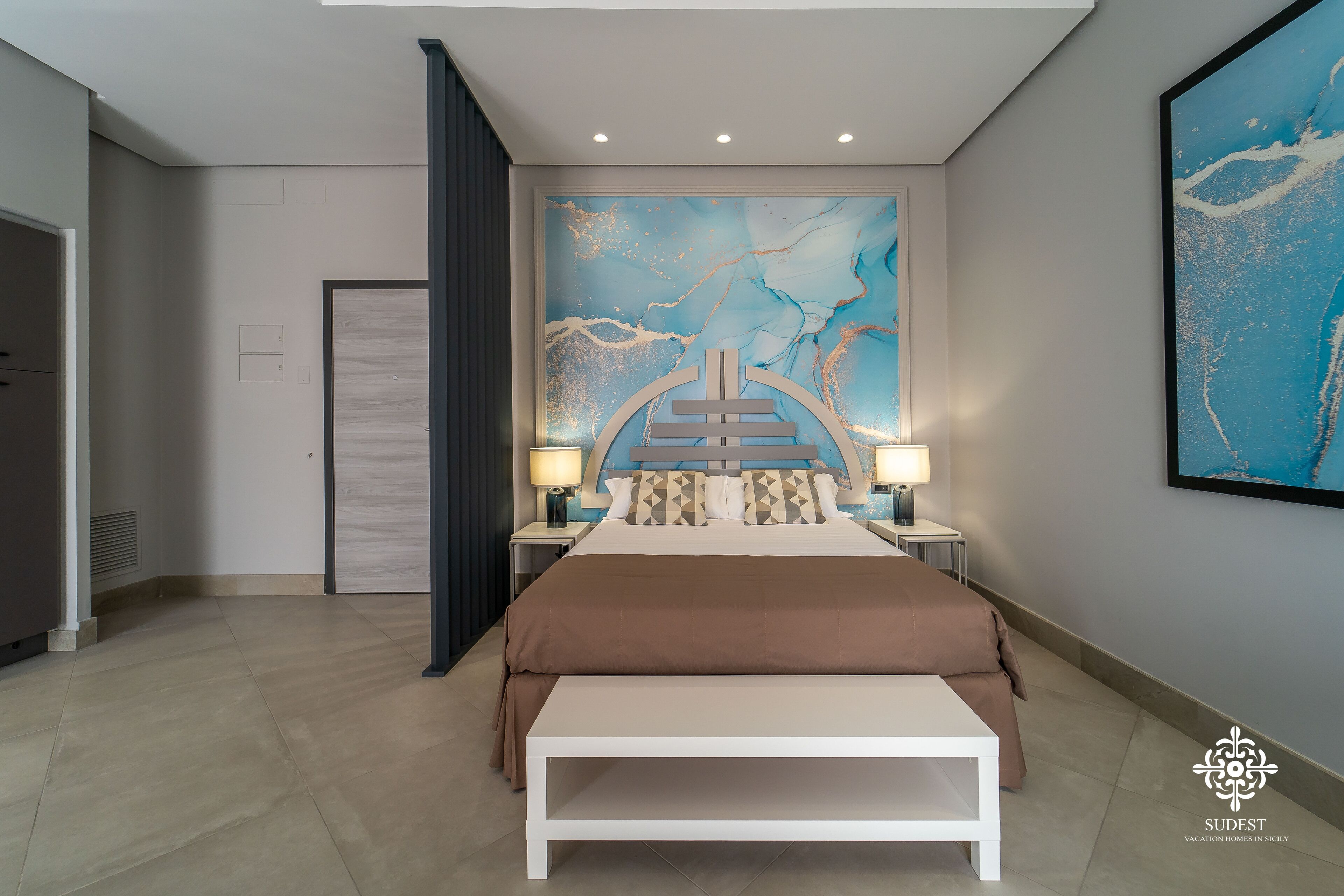 Une chambre à coucher moderne avec un grand lit, deux tables de chevet avec des lampes, une grande peinture abstraite au mur et un banc au pied du lit.