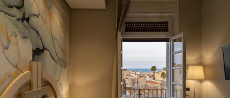 Habitación de hotel con cama, mesita de noche, lámpara, vista al mar y balcón con barandilla.