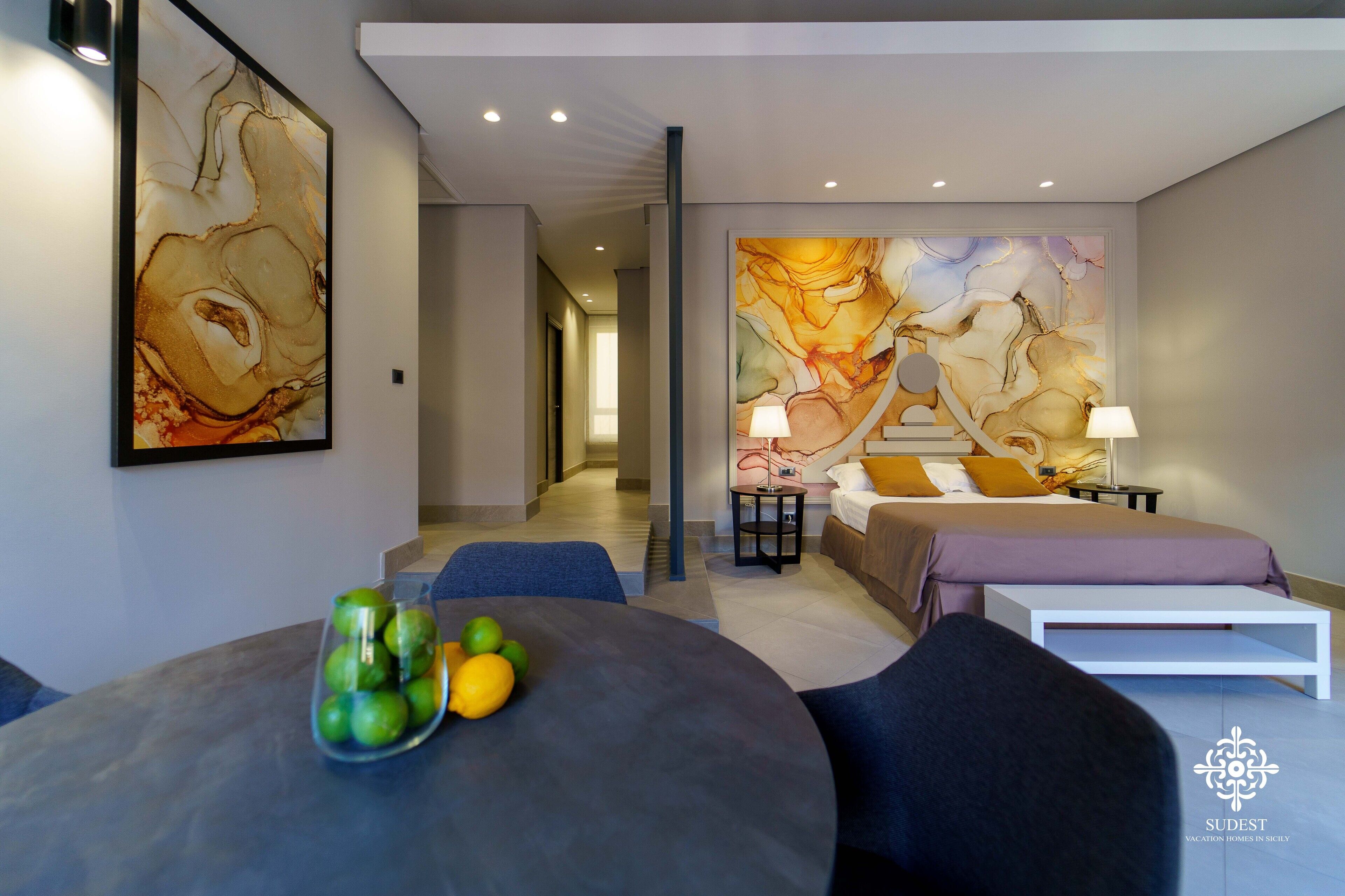 A modern hotel room with a large abstract painting on the wall, a bed with a purple bedspread, a small table with a fruit bowl, and a white nightstand with a lamp.