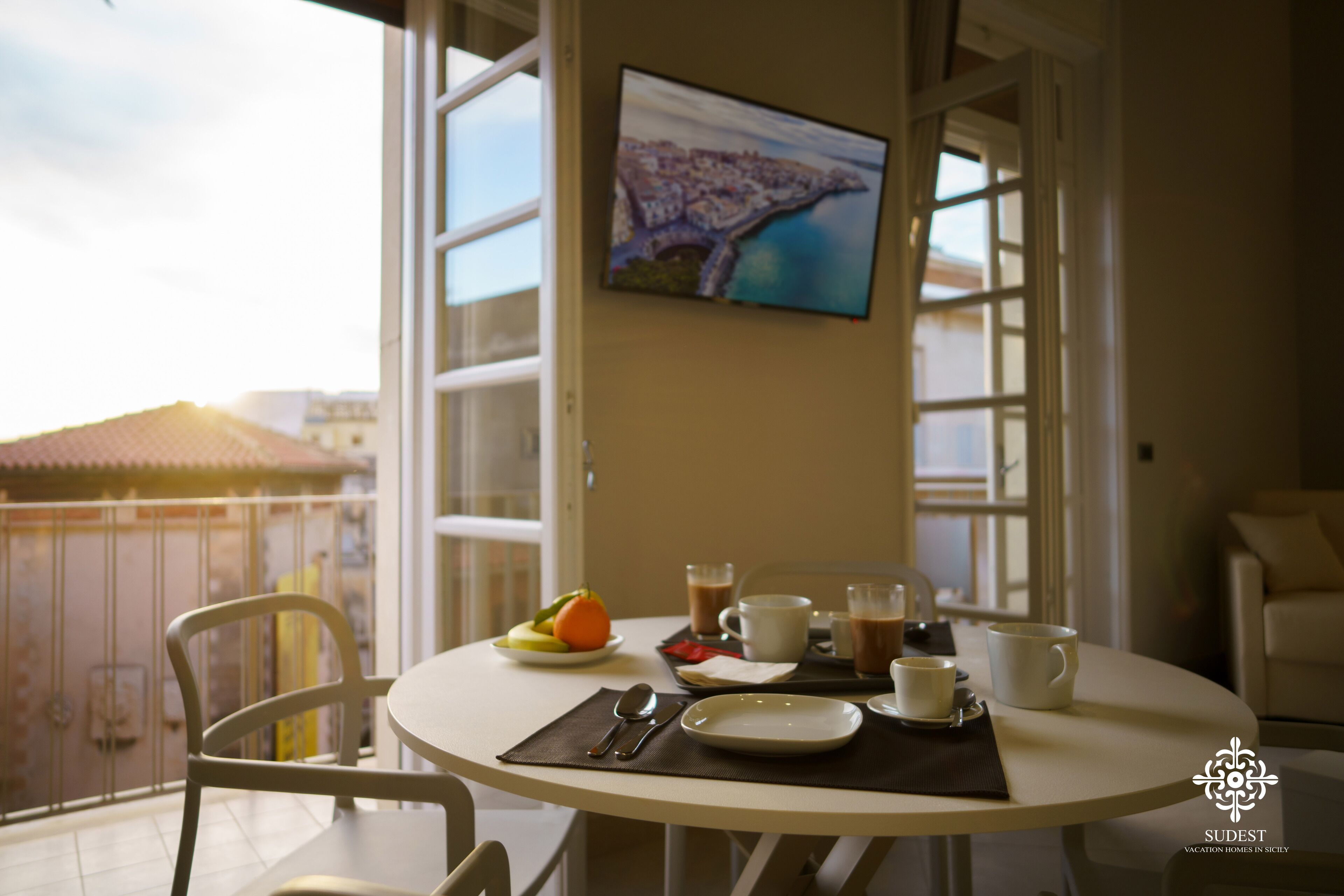 Une table à manger avec des aliments pour le petit-déjeuner, un téléviseur fixé au mur et un balcon offrant une vue sur les bâtiments.