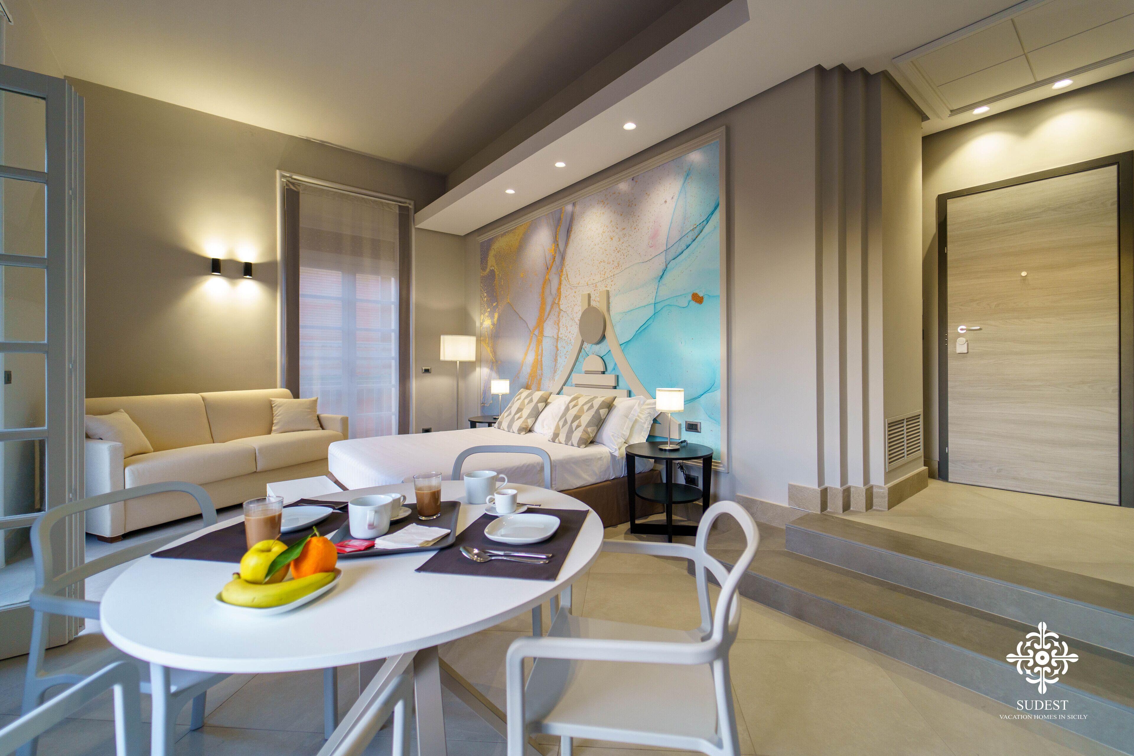 A modern hotel room with a bed, bedside tables, a dining area with a table and chairs, and a large abstract painting on the wall.
