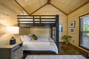 A wooden cabin bedroom with a bunk bed, a bedside table with a lamp, a dresser, and a potted plant.
