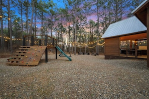 A wooden cabin with a deck and climbing wall, surrounded by a forest and string lights.