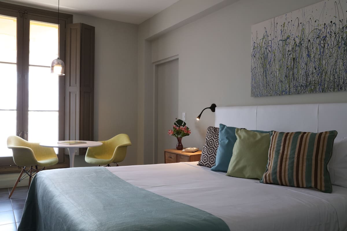 A hotel room with a bed, two yellow chairs, a small table, a nightstand with a lamp, and a painting on the wall.