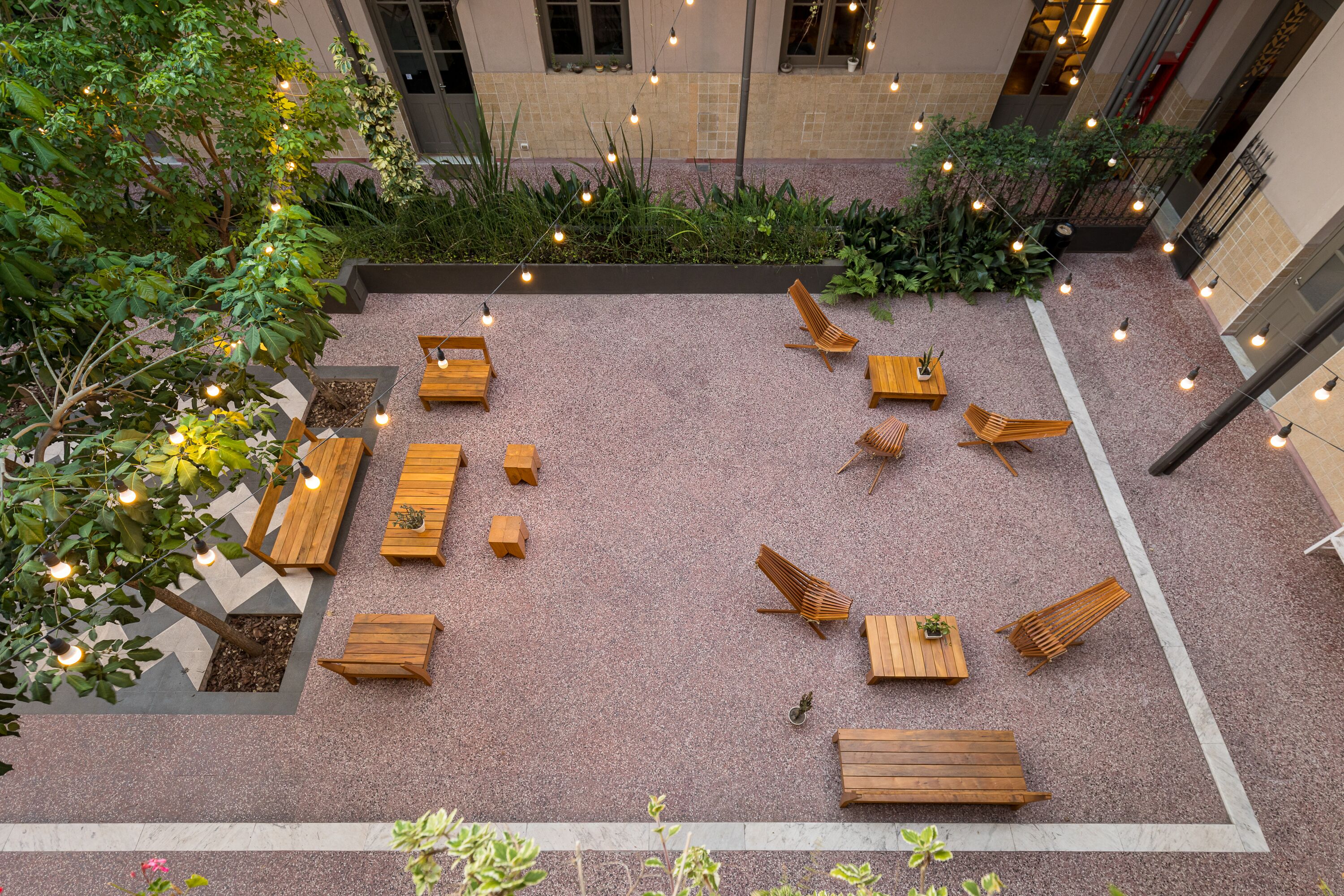 Un área de estar al aire libre con bancos y mesas de madera, rodeada de follaje y luces colgantes.