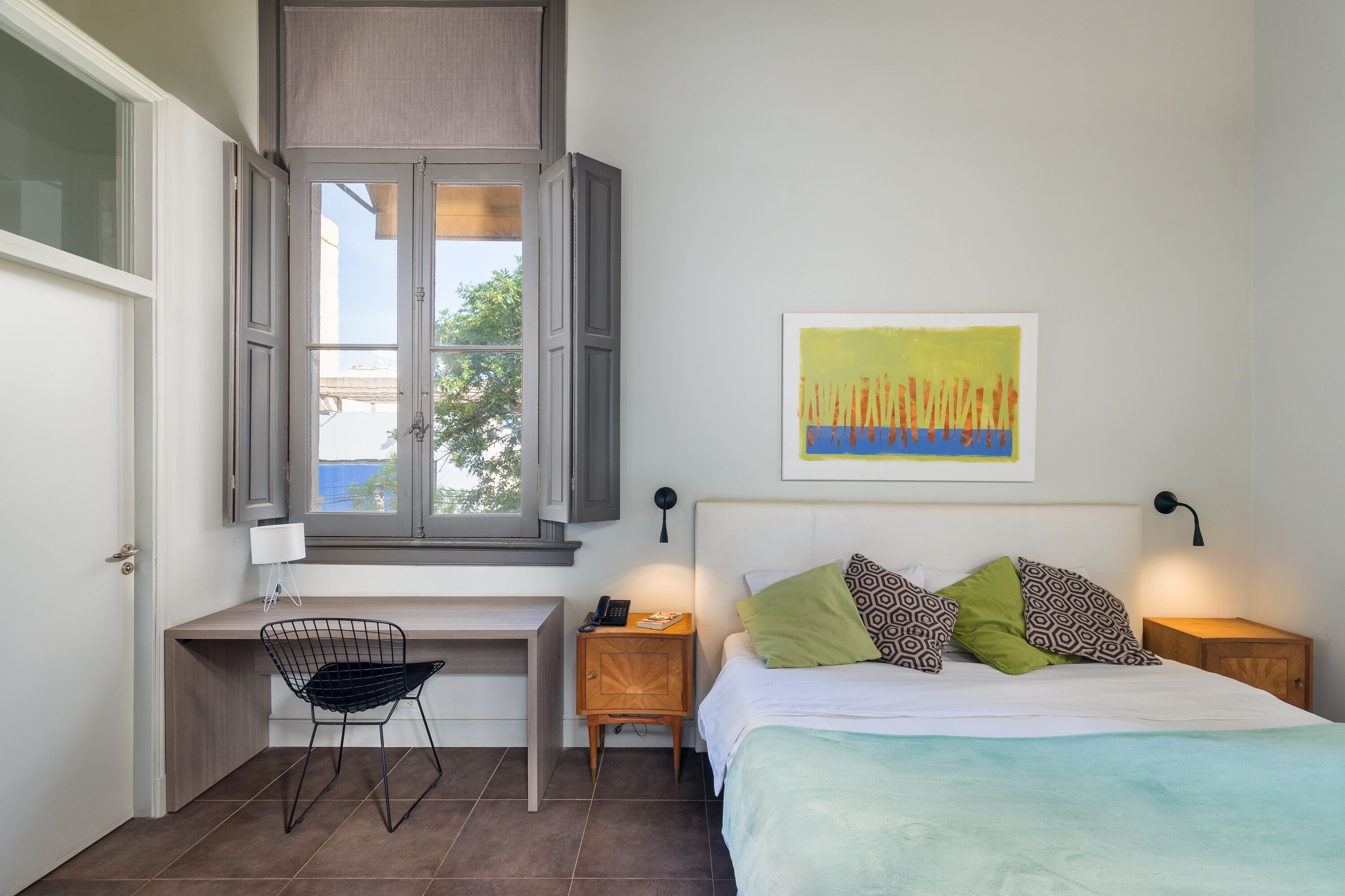 Un dormitorio con cama, escritorio, silla, ventana y una obra de arte en la pared.