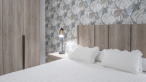 A bedroom with a bed, white bedding, a nightstand with a lamp, and a wall with a leaf pattern. - Cadiz Magnolia Apartment (Cádiz)