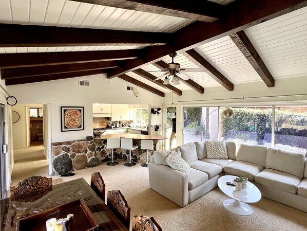 A spacious living area with a large sofa, a coffee table, and a stone accent wall. A kitchen is visible in the background, and there is a large window providing natural light.