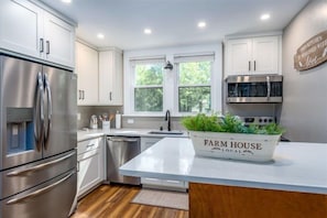 A modern kitchen with stainless steel appliances, a farmhouse-style island, and a window with a view of greenery.