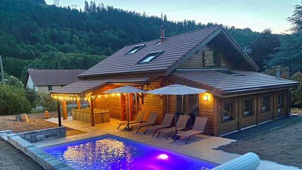 En stuga byggd av trä med en swimmingpool, utomhusmöbler och utsikt över en skogbeklädd sluttning.