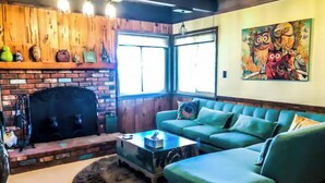 A cozy living room with a brick fireplace, a green sofa, and a colorful owl painting on the wall. - Relax with spectacular views @ Bear Dance Cabin (Running Springs)