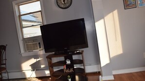 A living room with a flat-screen TV on a wooden stand, a window with blinds, a wall clock, and framed pictures.