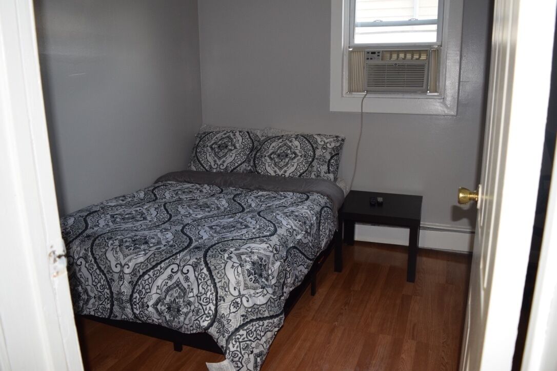 A bedroom with a bed, a nightstand, a window with an air conditioner, and a door.