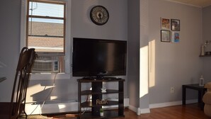 A living room with a flat-screen TV on a black stand, a clock on the wall, and a window with blinds.