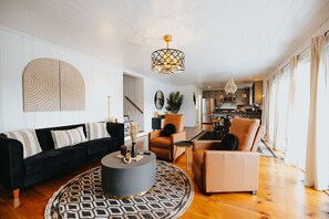 A modern living room with a black sofa, a round coffee table, and a beige armchair. There is a kitchen in the background with stainless steel appliances.