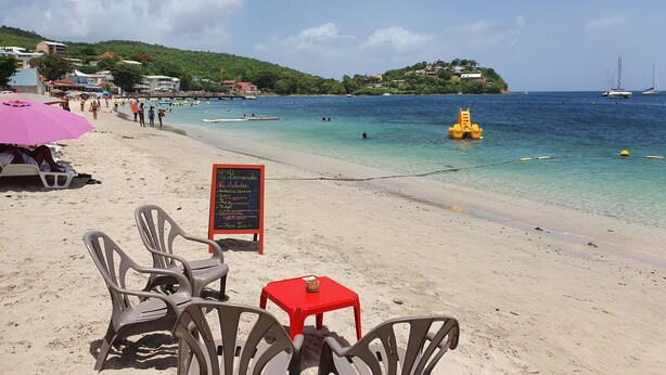 A beachside restaurant with outdoor seating, a menu board, and a view of the sea and boats.