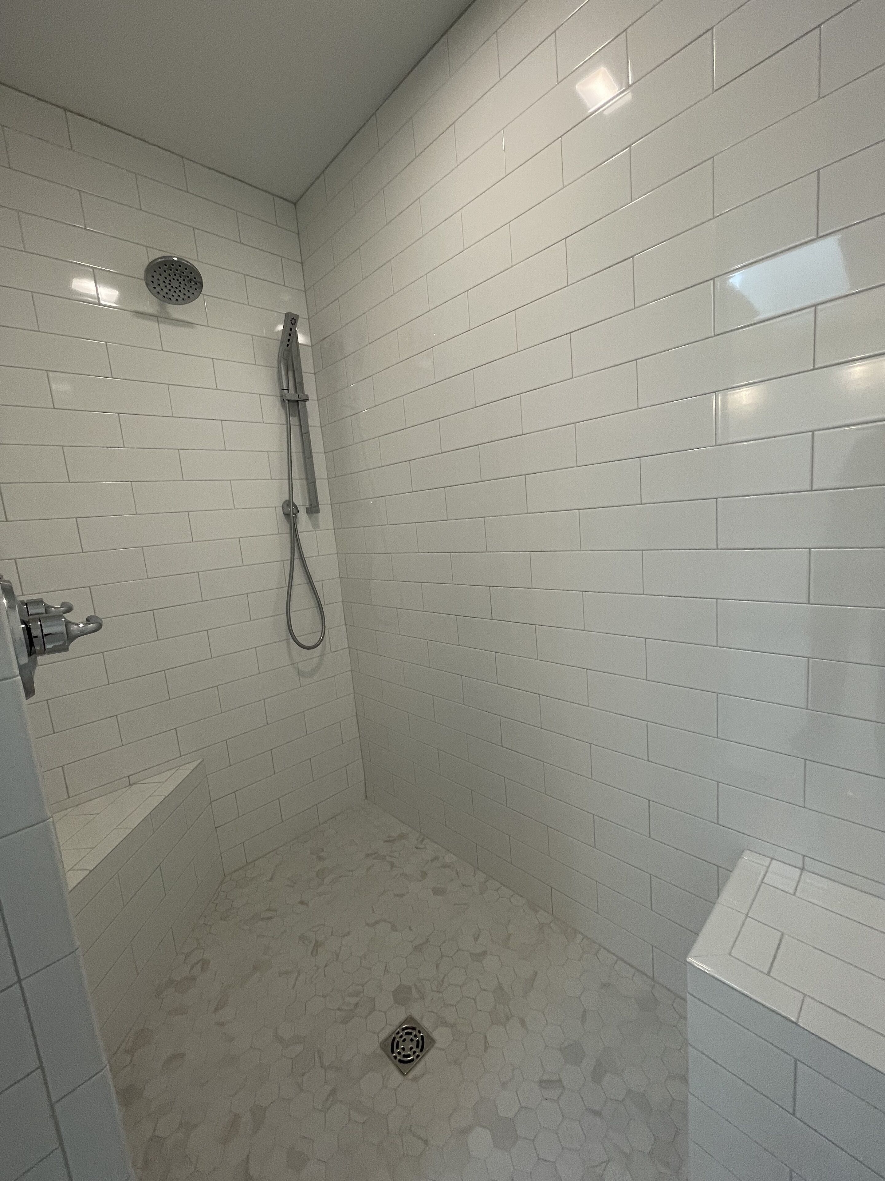 Une petite salle de douche aux murs recouverts de carreaux blancs, équipée d’un pommeau de douche et d’un flexible de douche.