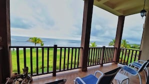 A covered balcony with a view of the ocean, a grassy area, and a few chairs. - Suite San Juan 125 Gran Pacifica Resort (Villa El Carmen)
