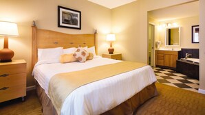 Habitación de hotel con una cama grande, dos lámparas de noche, cabecera de madera, un cuadro enmarcado en la pared, baño con lavamanos y espejo, y piso a cuadros.