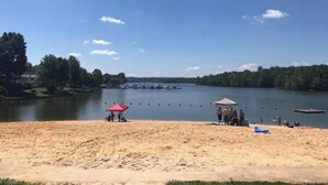 A sandy beach with a red umbrella and a small structure, likely a lifeguard station, near a calm lake surrounded by trees.