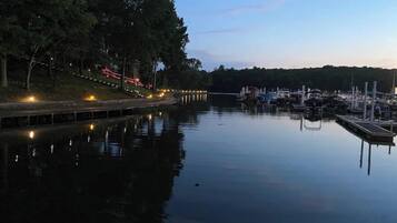 Un puerto deportivo tranquilo al anochecer, con barcos amarrados, un sendero iluminado y un fondo arbolado.