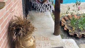 Una hamaca bajo una pérgola de madera, un muro de ladrillo, una puerta y un suelo de baldosas con una planta en maceta y un jarrón decorativo.