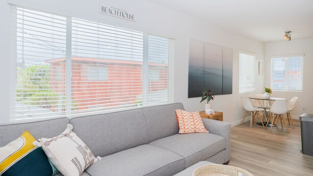A modern living room with a grey sofa, a dining table with chairs, and large windows with blinds.