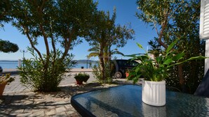 Ein Tisch mit Glasplatte, mit einer weißen Vase und umgeben von Topfpflanzen, in einer sonnigen Außenanlage mit Blick auf das Meer und einem parkenden Auto.
