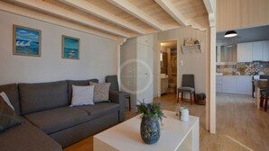 Ein modernes Wohnzimmer mit einer grauen Couch, einem weißen Couchtisch und einer Vase mit Blumen. Es gibt eine Küchenzeile mit weißen Schränken und einer gefliesten Spritzschutzwand.