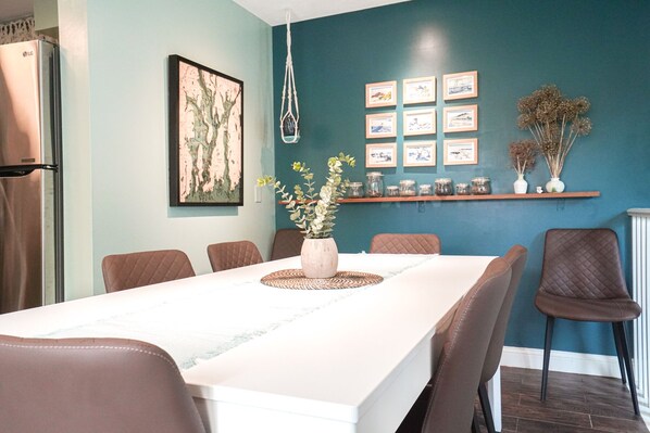A dining area with a long table, six chairs, a vase with flowers, and a wall with framed pictures and shelves with jars.