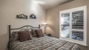 Une chambre à coucher comprenant un lit, une table de chevet, une lampe, un élément de décoration murale et une fenêtre avec des stores.