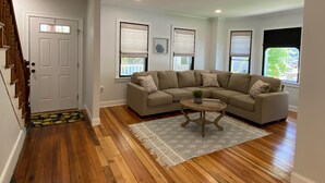 A living room with a beige sectional sofa, a round coffee table, and a patterned rug. There are three windows with blinds, a white door, and a staircase with wooden steps.