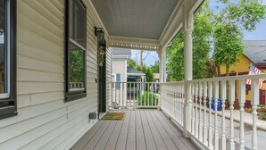 A covered porch with a white railing, a view of the street, and a yellow house with an American flag in the background.