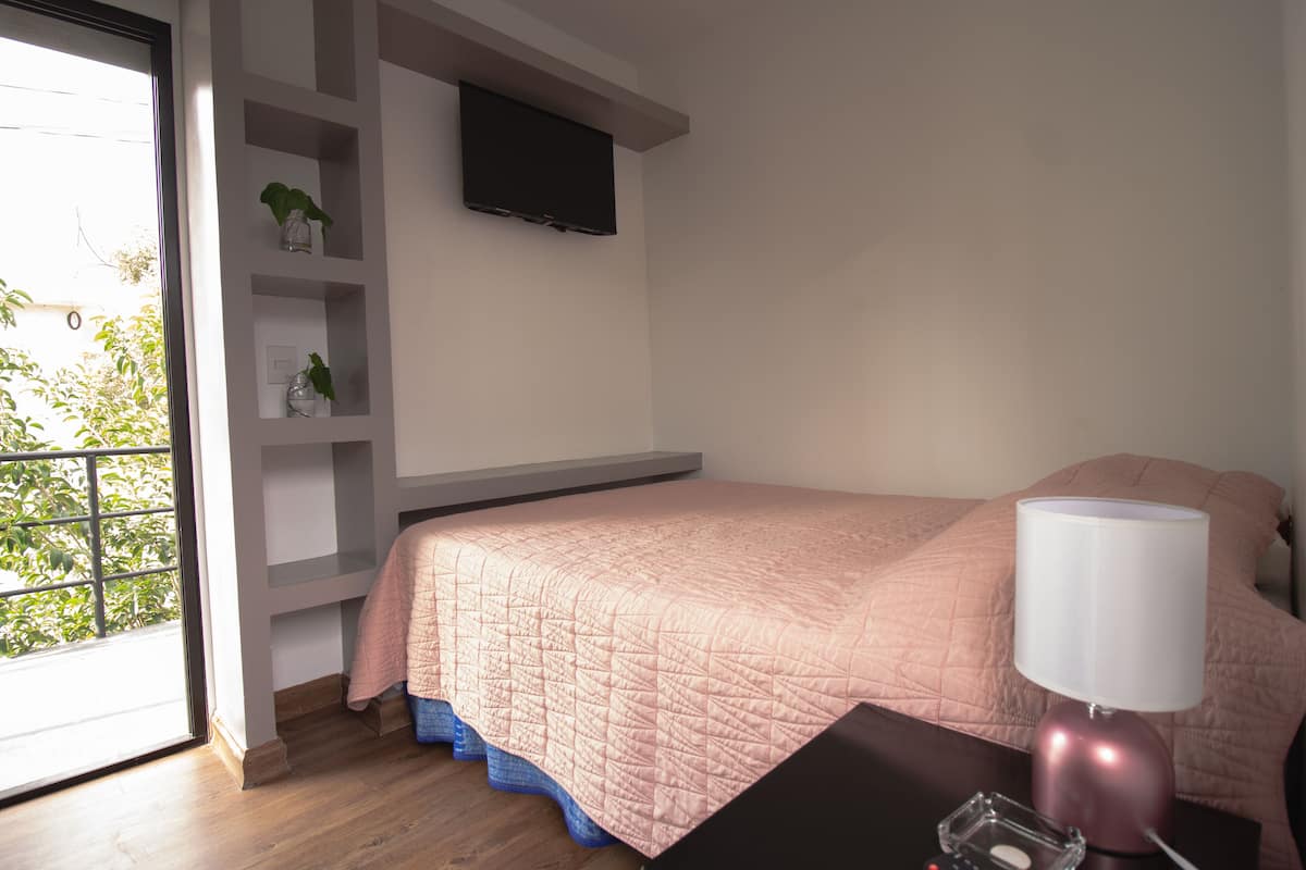 Un dormitorio con cama, una mesita de noche con lámpara, un televisor montado en la pared y una ventana con vistas a zonas verdes.