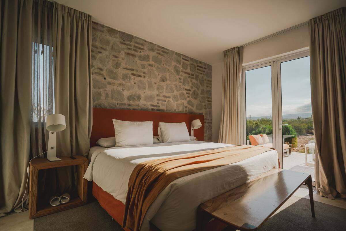 A bedroom with a stone wall, a large bed with white bedding, a wooden bench, and a view of the outdoors.