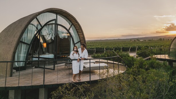 A couple standing on a wooden deck in front of a geodesic dome structure with a glass wall, overlooking a vineyard. - Chozos Resort (Ugarteche)