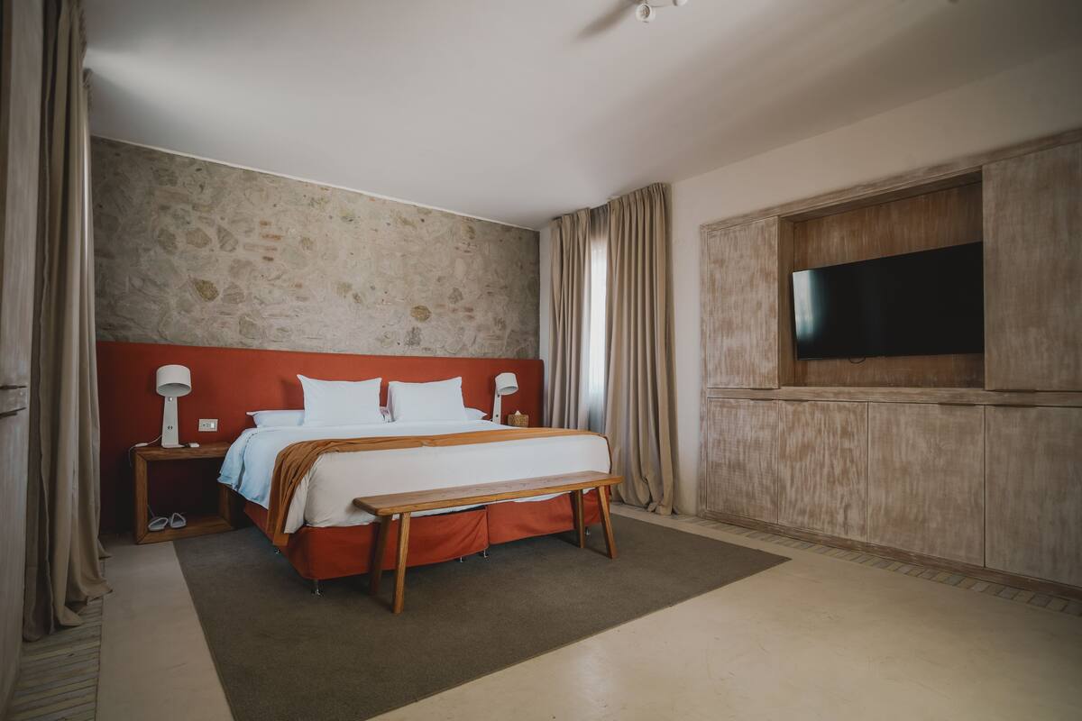A hotel room with a large bed, a wooden bench, a TV mounted on the wall, and a stone-textured wall.