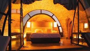 A cozy, circular room with a bed centered under a large arched window, a small table, and a chair. - Chozos Resort (Ugarteche)