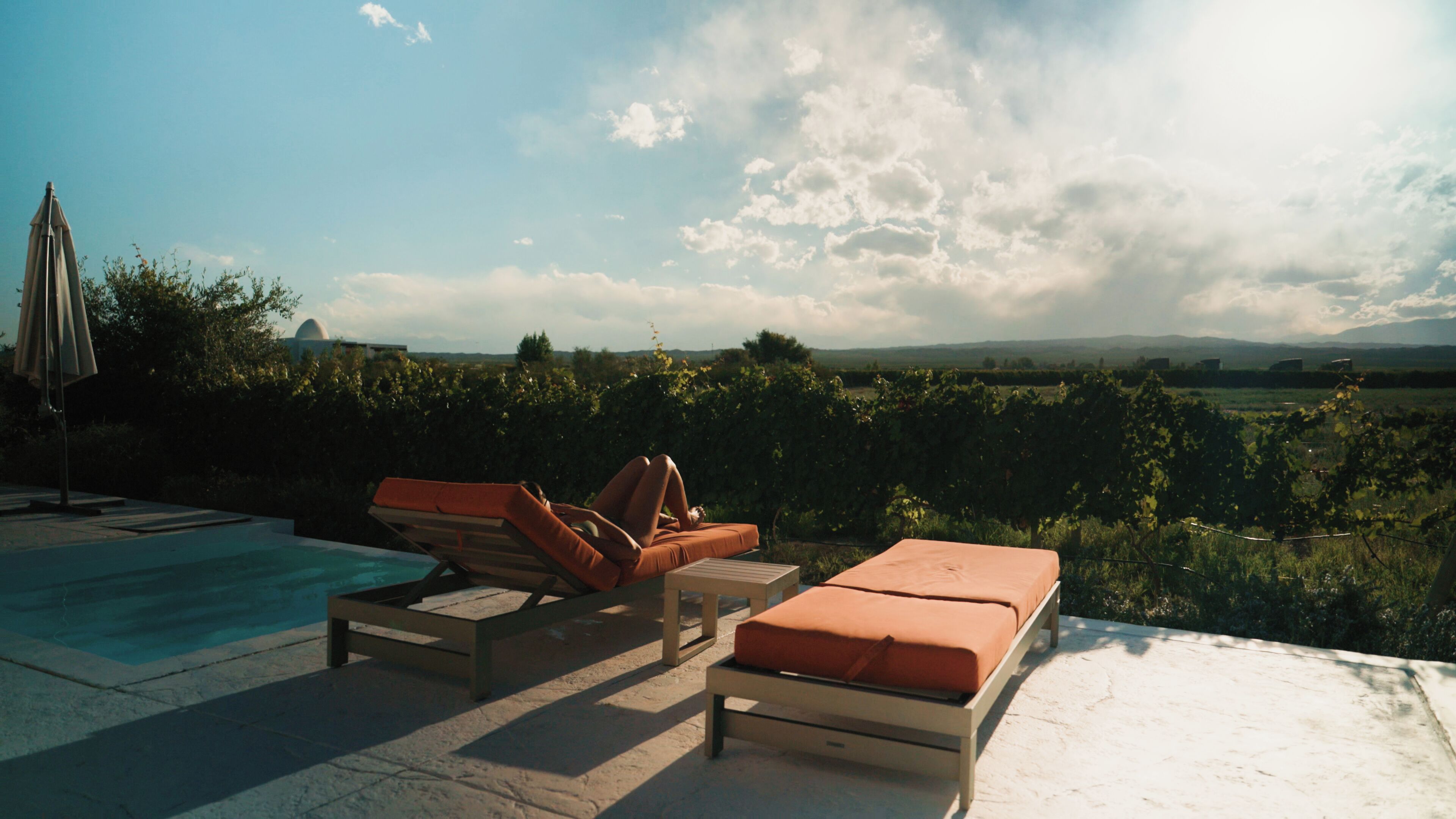 Zona junto a la piscina con sillones de descanso y una pequeña mesa, con vistas a un viñedo y colinas lejanas.